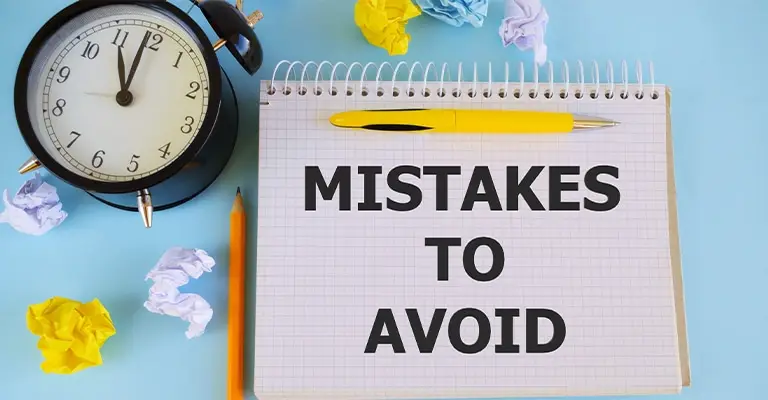 Flat lay image featuring a spiral notebook with the bold text “MISTAKES TO AVOID” on a gridded page. A yellow pen rests on top of the notebook, surrounded by a black alarm clock, pencil, and crumpled paper balls on a light blue background, suggesting brainstorming or editing errors.