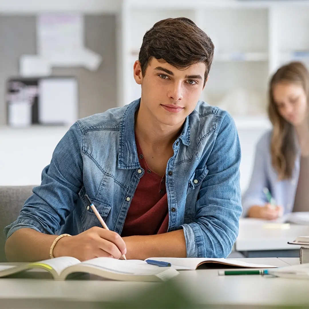 A young man in a denim shirt sits at a desk, writing in a notebook, with a focused expression.