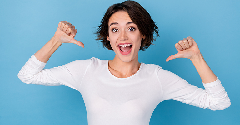Excited young woman in a white shirt pointing at herself with both thumbs against a bright blue background.