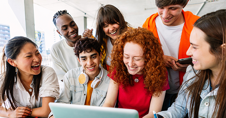 Group of diverse students gathered around a laptop, laughing and collaborating, representing teamwork, friendship, and positive student engagement.