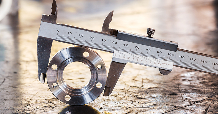 A precision caliper measures the outer diameter of a shiny metal part with bolt holes, resting on a scratched industrial work surface.