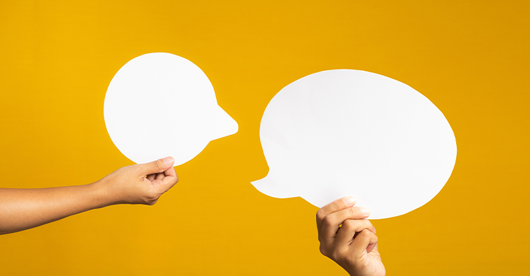 Two hands hold up white paper speech bubbles against a bright yellow background, symbolizing open communication and meaningful conversation.