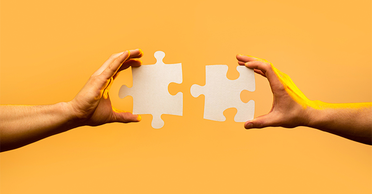 Two hands holding white puzzle pieces on an orange background, symbolizing collaboration, teamwork, or problem-solving.