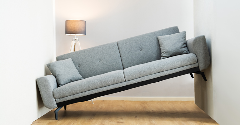 Gray sofa stuck and tilted between two white walls on a wooden floor with a lit floor lamp in the background, illustrating a tight space challenge.
