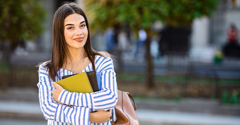 A young woman with long dark hair stands outdoors holding a green notebook and smiling confidently. She wears a striped shirt and carries a tan backpack, with trees and a blurred campus setting in the background.