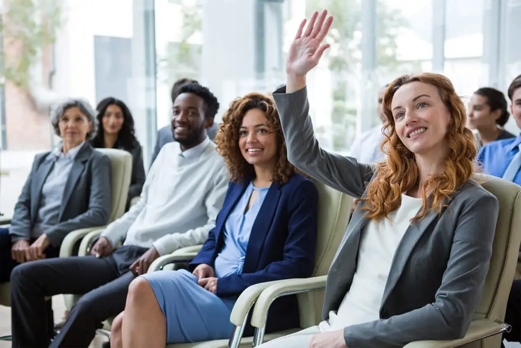 Digital Footprint Audit image of professionals seated in a meeting or training room with one woman raising her hand to participate. The scene suggests reviewing workplace presence and reputation in a group setting.