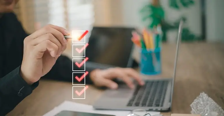 A person’s hand checks off items on a vertical checklist overlay while working on a laptop at a desk with a cup of colored pencils in the background. The image suggests completing tasks or reviewing a process, with multiple red check marks indicating progress.