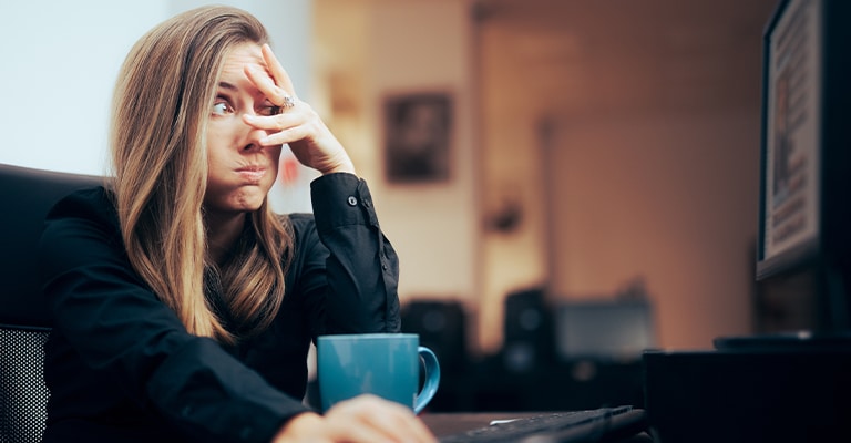 A frustrated woman sits at her desk, staring at a computer screen with her hand partly covering her face and puffed cheeks. A blue coffee mug sits nearby, suggesting fatigue or overwhelm during a long workday.