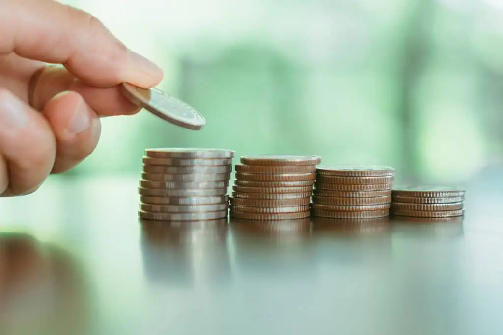 A close up hand places a coin onto stacks of coins lined up from tallest to shortest on a reflective table. The image suggests saving money or building wealth through gradual financial growth.