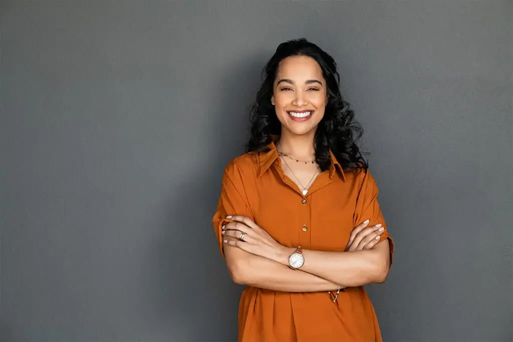 A smiling woman with curly dark hair stands with her arms crossed against a plain gray background. She wears a rust orange button up shirt with a wristwatch and simple jewelry for a confident professional portrait look.