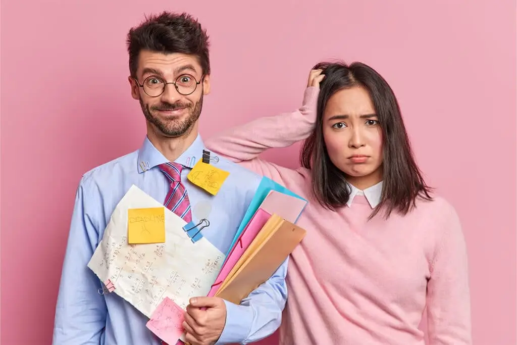 A man in glasses and a tie holds a stack of papers folders and sticky notes while smiling in front of a pink background. Next to him a woman in a pink sweater looks worried and scratches her head while holding colorful file folders suggesting workplace stress and looming deadlines.