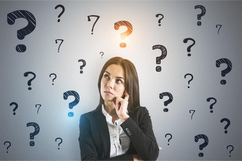 A woman in a business suit looks up thoughtfully with her hand resting on her chin. Large question marks surround her on a gray background including one highlighted above her head to show uncertainty or problem solving.
