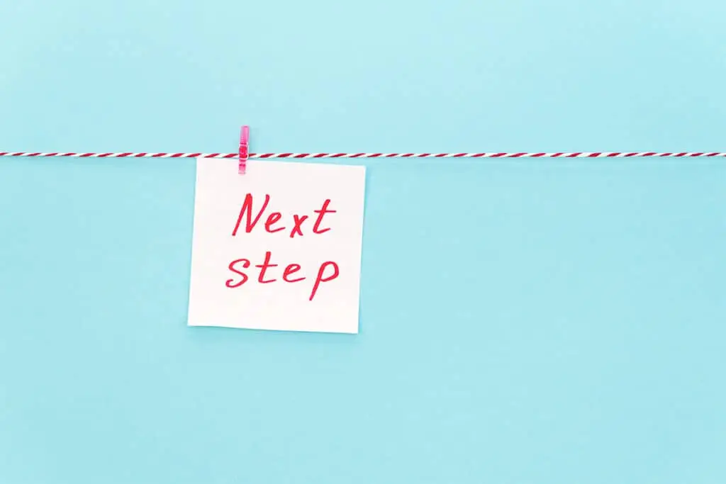 A small note card clipped to a red and white string reads Next step in red handwriting against a light blue background. The simple layout highlights taking the next step or moving forward in a plan.
