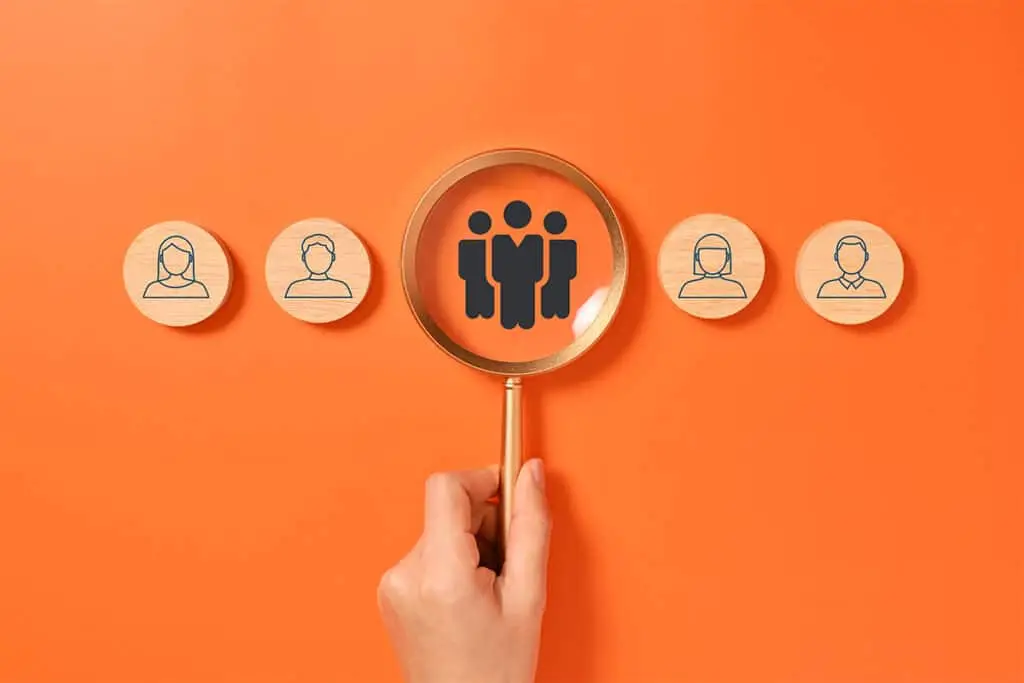 A hand holds a magnifying glass over a group icon on an orange background, with small wooden profile tokens lined up on both sides. The image suggests identifying an audience or community to help build a positive professional online reputation.