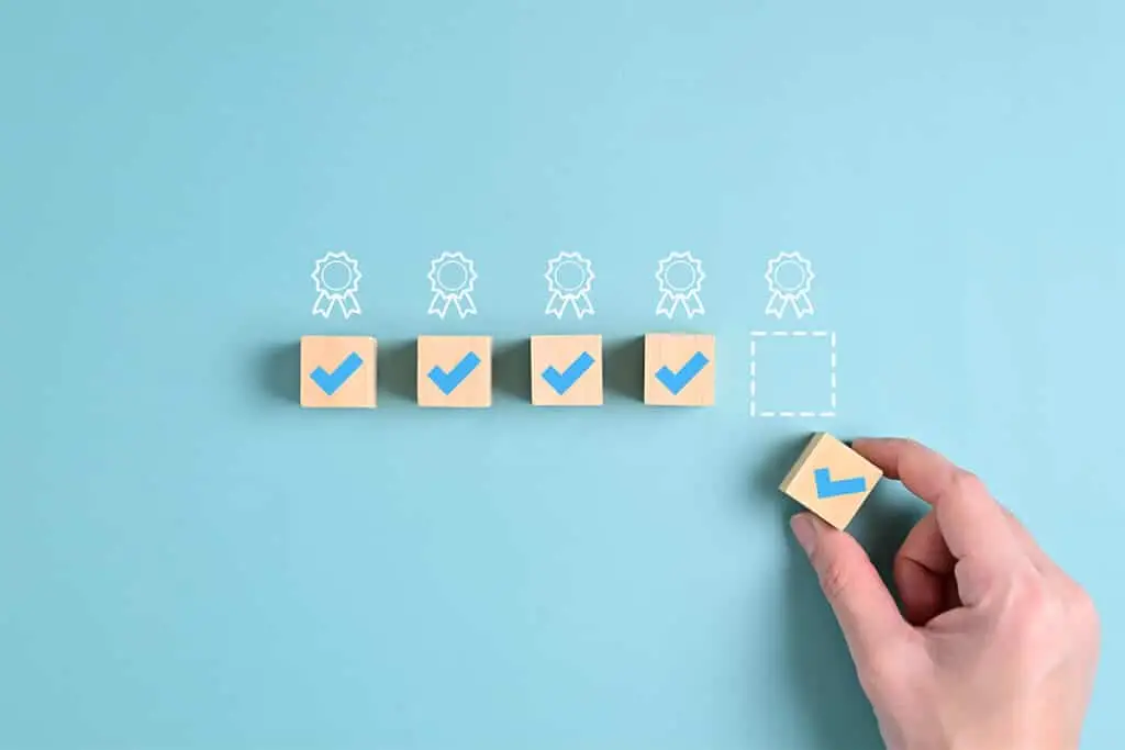 A hand places a wooden block with a blue check mark into an empty dotted square, completing a row of checked boxes on a light blue background. The image suggests meeting quality standards and consistently delivering results that support a positive professional online reputation.