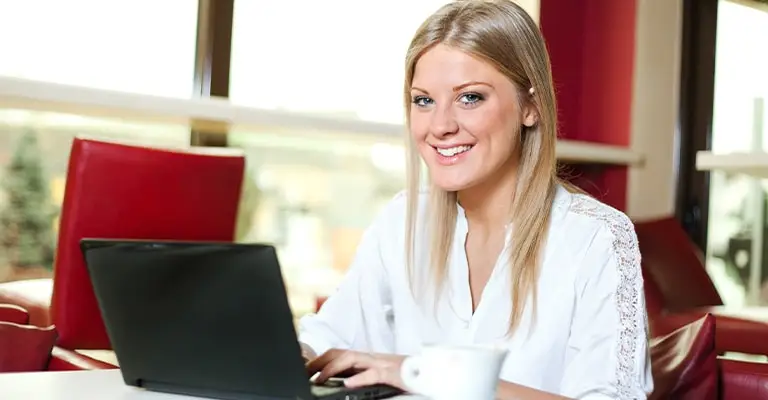 A smiling woman uses a laptop in a bright cafe. The scene supports advice to protect your online reputation by managing what appears about you online.