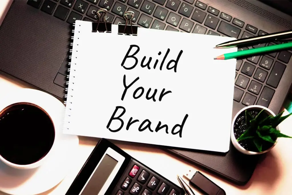 A notebook on a laptop keyboard shows the handwritten words Build Your Brand, surrounded by a coffee cup, pens, a calculator, and a small plant on a desk. The image represents planning and marketing work to Build a Personal Brand and strengthen a positive professional online reputation.