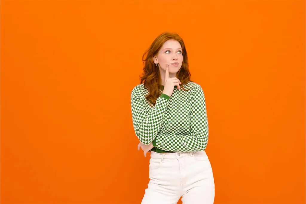 A person stands against a bright orange background with a thoughtful expression, one hand on their chin and the other arm crossed. The image represents considering your identity and messaging as you Build a Personal Brand.
