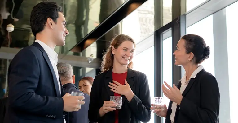 Three professionals in business attire talk and smile while holding drinks at a networking event. Their conversation illustrates how building professional relationships can strengthen a professional brand.