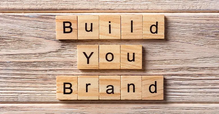 Wooden letter tiles spell "Build You Brand" on a rustic wood surface. The missing letter in "Your" draws attention to the process of learning how to build a professional brand.