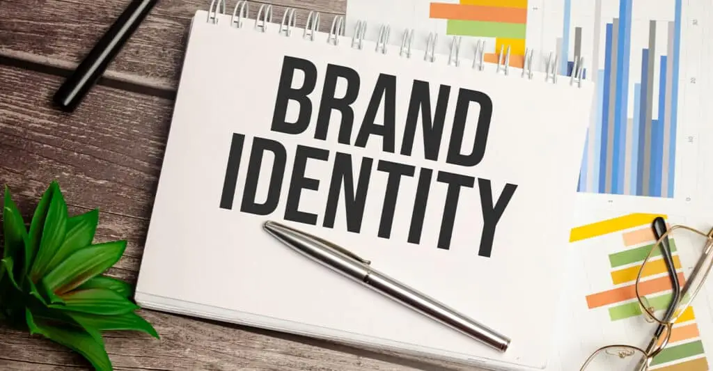 A spiral notebook on a desk reads BRAND IDENTITY in bold letters, with a pen laid across the page and colorful bar charts and graphs in the background. Visual for develop your personal brand by focusing on brand identity and how it connects to planning and results.