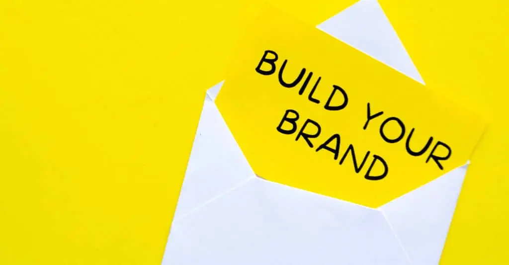 A white envelope on a bright yellow background holds a card that reads BUILD YOUR BRAND in bold black letters. Simple visual for develop your personal brand and sharing your message through clear branded communication.