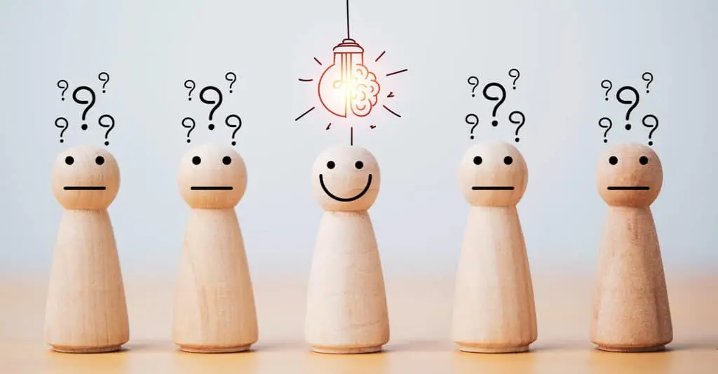 Five wooden peg figures stand in a row on a light background, with question marks above four of them and a smiling figure in the center under a hanging light bulb with a brain icon. Concept image for develop your personal brand by finding clarity and a standout idea while others feel uncertain.