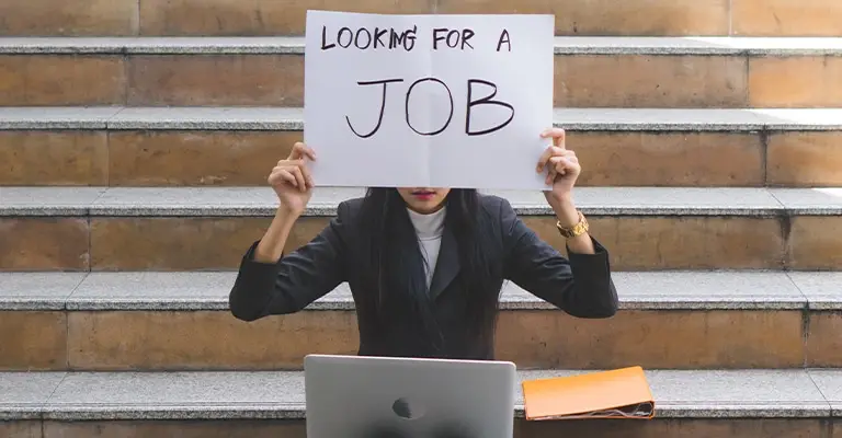 A person sits on outdoor steps with a laptop and holds a handwritten sign that says "LOOKING FOR A JOB" in front of their face. The scene illustrates the uncertainty and effort behind entry-level job search tips for new applicants.