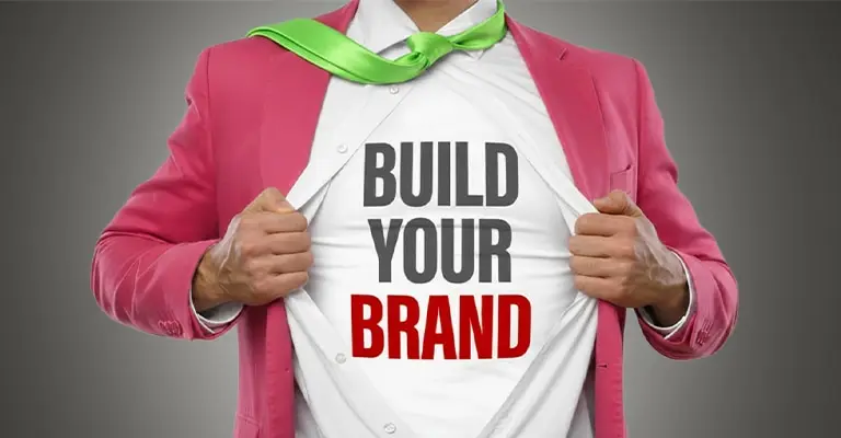 Person in a bright pink suit and green tie pulls open a white shirt to reveal the words "BUILD YOUR BRAND." The bold superhero style visual represents building your brand on LinkedIn.
