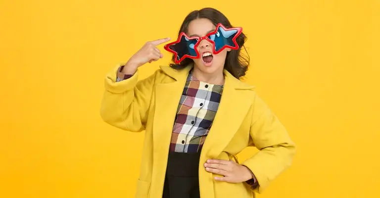 Woman in a bright yellow coat points toward oversized red and blue star shaped sunglasses while posing against a yellow background. Her playful confident expression reflects standing out while you build your brand on LinkedIn.