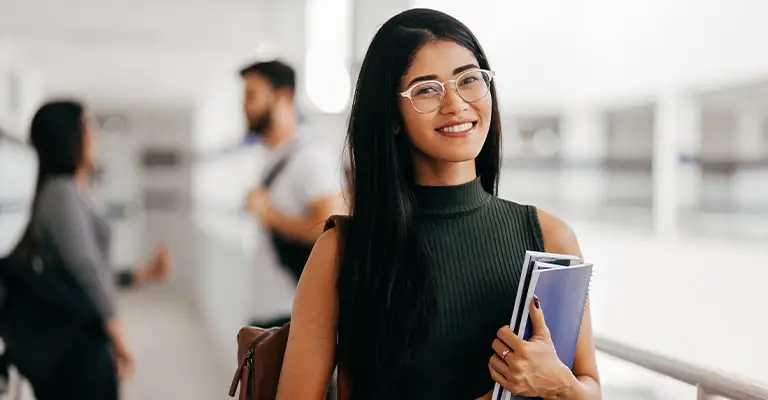 A smiling student wearing glasses holds notebooks in a bright campus hallway while other students are blurred behind her. The polished portrait fits a personal branding for students topic by showing confidence and approachability.