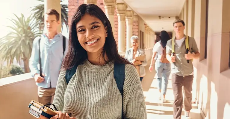 A smiling college student with a backpack holds notebooks in a sunlit campus walkway while other students pass behind her. The approachable portrait fits personal branding for college students by showing confidence and connection on campus.