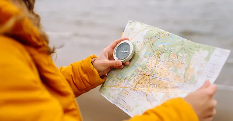 A person in a yellow jacket holds a paper map and compass while standing near the water. The close up focuses on navigation and planning during an outdoor trip.