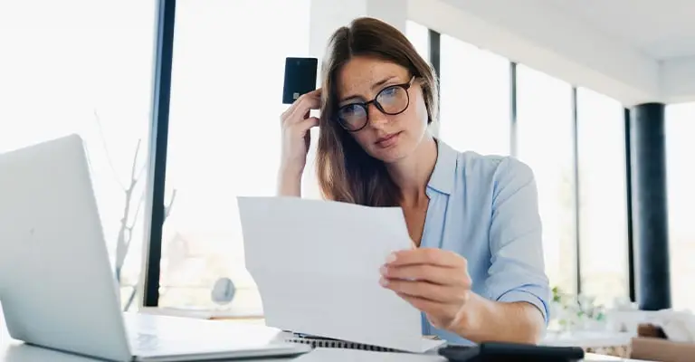 A woman with glasses reviews a document at a desk with a laptop nearby while holding a black card near her head. Her focused expression suggests she is making a careful financial or professional decision.