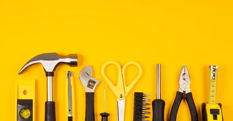 Hand tools are arranged in a neat row on a bright yellow background including a level. hammer. utility knife. wrench. scissors. screwdriver. bits. pliers. and tape measure.