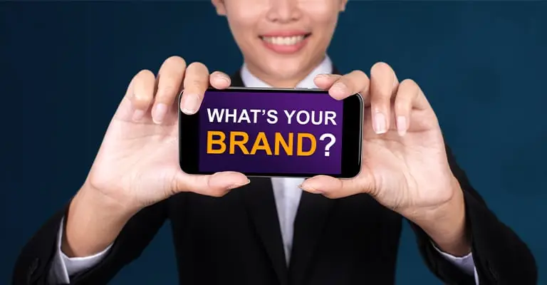 A person in a black blazer holds a phone toward the camera with the screen text “WHAT’S YOUR BRAND?”. The direct question and business style make it useful for a personal branding for students article about defining how others see you.