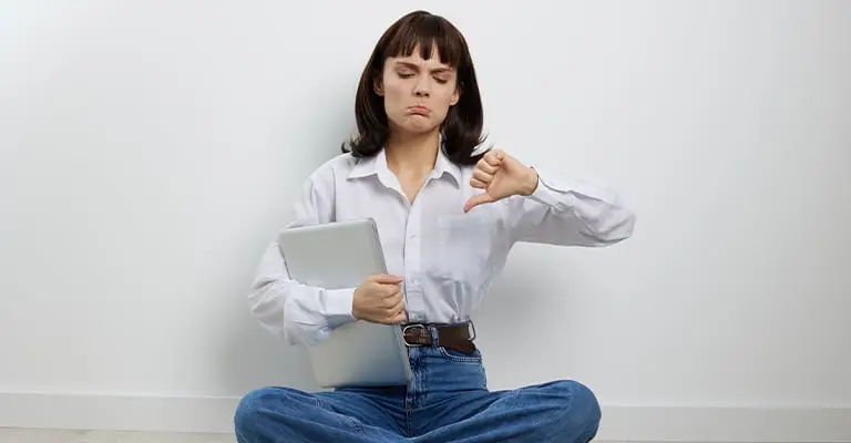 A young woman sits cross legged on the floor holding a silver laptop and making a thumbs down gesture. Her unhappy expression fits a personal branding for students article about mistakes to avoid when building an online presence.