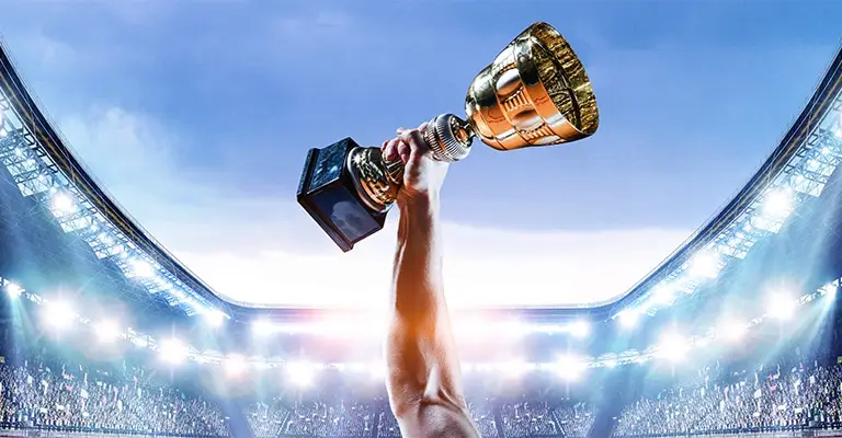 A raised arm holds a large gold trophy above a brightly lit stadium filled with spectators. The winning moment represents achievement recognition and success through early career advancement.