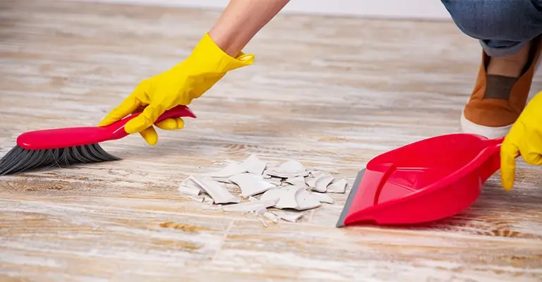 A person wearing yellow rubber gloves sweeps broken white pieces into a red dustpan on a wooden floor. The cleanup suggests addressing mistakes and removing obstacles as part of early career advancement.