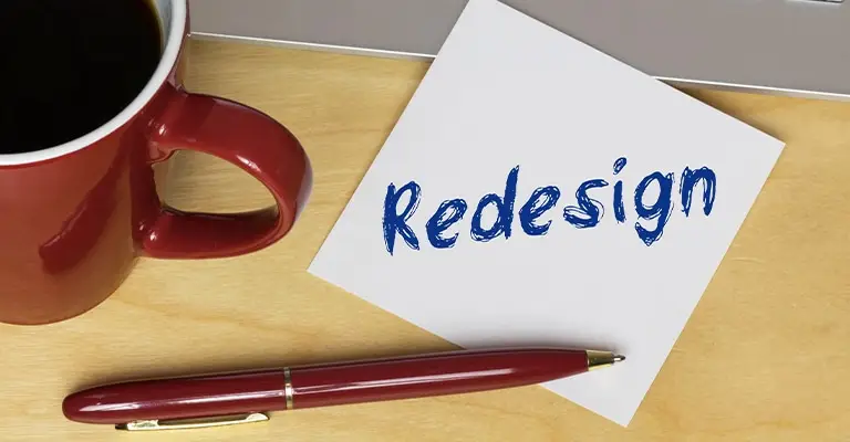 A white note marked "Redesign" sits beside a red coffee mug and matching pen on a wooden desk. The workspace represents reassessing goals and redesigning a professional path for early career advancement.