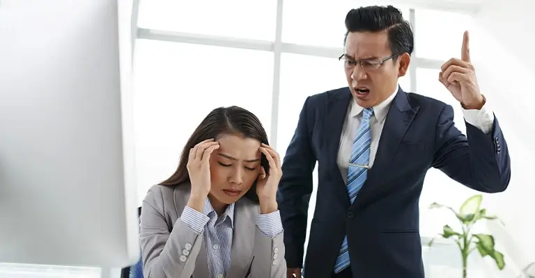 A male manager points upward while angrily speaking to a seated female employee who holds her head in distress at a computer. The tense workplace interaction highlights the communication and leadership challenges that can affect early career advancement.