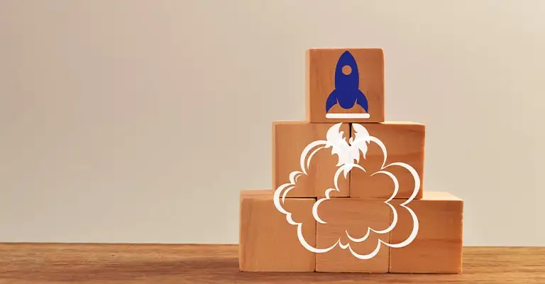 A blue rocket launches from the top of stacked wooden blocks as white smoke curls around the base. The upward movement symbolizes momentum growth and new opportunities in early career advancement.