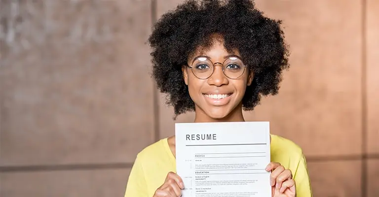 A smiling college student with glasses holds a document titled “RESUME” toward the camera. The portrait highlights resume preparation for a professional website for college students.
