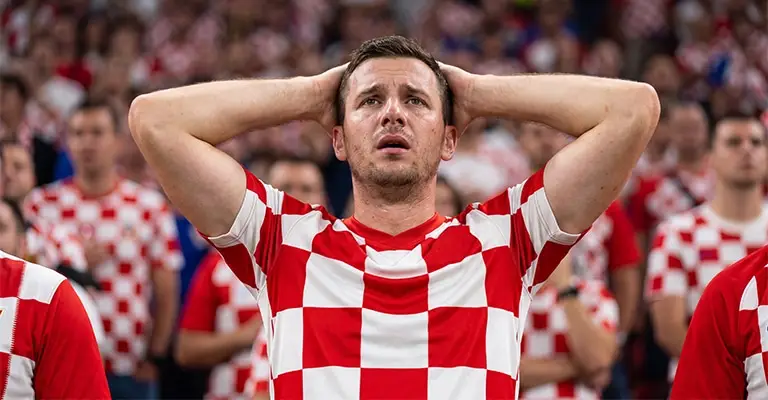 Croatian soccer fan in a red and white checkered jersey holds his hands on his head with a worried expression in a crowded stadium. The frustrated reaction can represent the regret people feel before learning how to avoid LinkedIn mistakes.