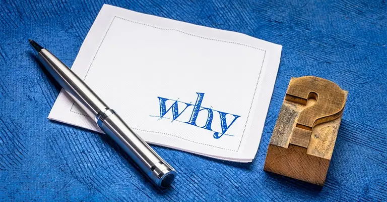 A silver pen rests beside a white card displaying the handwritten word “Why” on a textured blue surface. A wooden question mark reinforces the importance of explaining purpose and value during LinkedIn profile optimization.