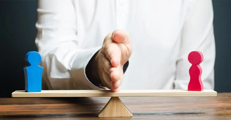 A person holds one hand upright between blue and pink wooden figures balanced on opposite ends of a seesaw. The scene represents defining professional boundaries and separating audiences during LinkedIn profile optimization.