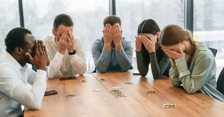Five coworkers sit around a conference table with their faces covered during a card game. Their visible frustration suggests the confusion and missed opportunities that LinkedIn profile optimization can help prevent.