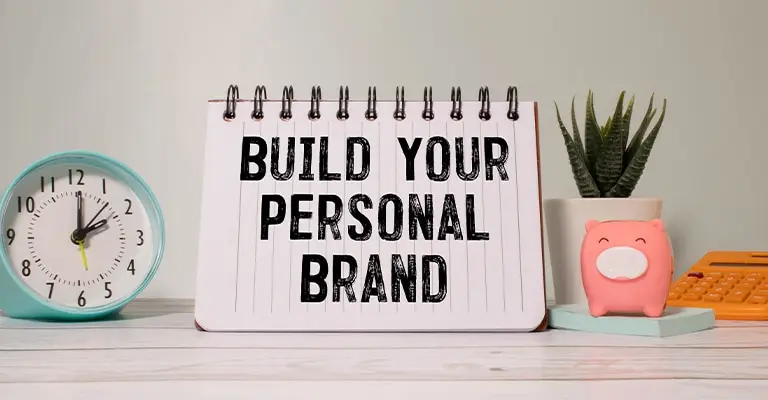 Spiral notebook on a desk displays the text “BUILD YOUR PERSONAL BRAND.” A blue clock, small potted plant, pig shaped container, and orange calculator frame the notebook like a workspace for planning a personal branding strategy.
