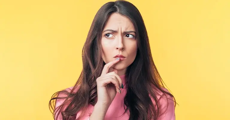 Thoughtful woman in a pink top rests a finger near her lips while looking to the side against a yellow background. Her uncertain expression reflects the questions that can arise when trying to optimize your social media.