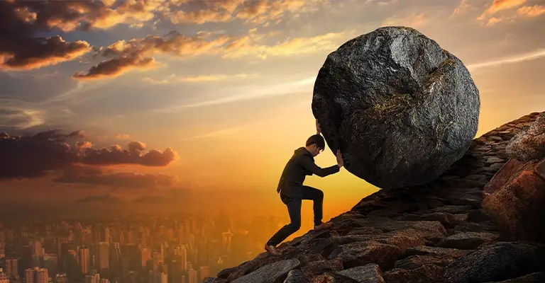 A person in business attire strains to push a massive boulder uphill on a rocky slope at sunset with a city skyline in the background. The dramatic scene symbolizes the value of personal branding by representing the persistence and effort required to build a strong professional reputation and stand out over time.